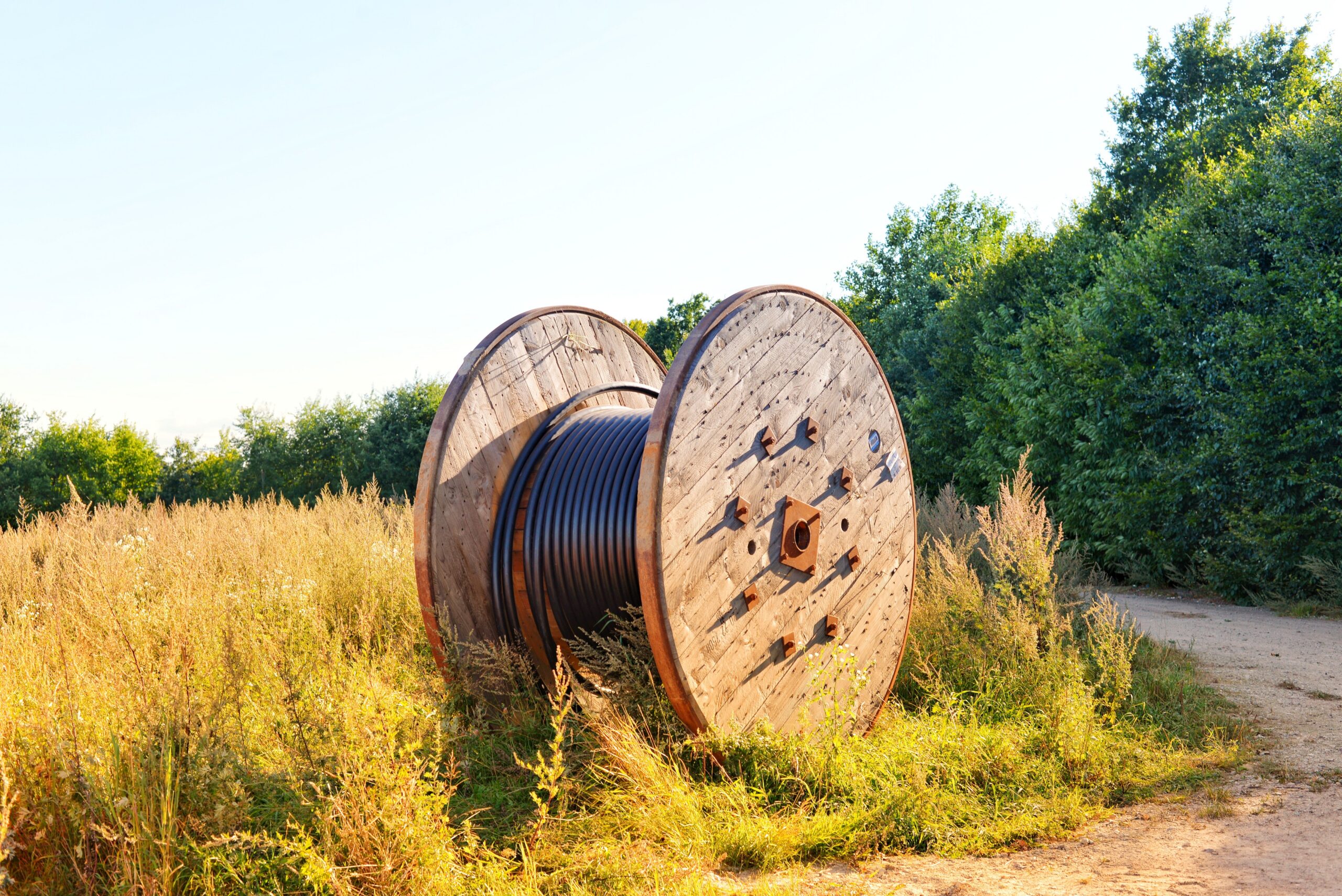 Wooden cable reel with coiled wires in a sunlit field. Ideal infrastructure stock photo.